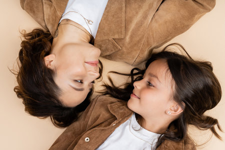 top view of mom and daughter in trendy autumn jackets smiling at each other on beige backgroundの写真素材
