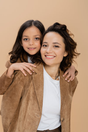 brunette girl hugging shoulders of happy mom in brown suede jacket isolated on beigeの写真素材