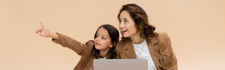 trendy girl pointing with finger near laptop and amazed mom isolated on beige, bannerの写真素材