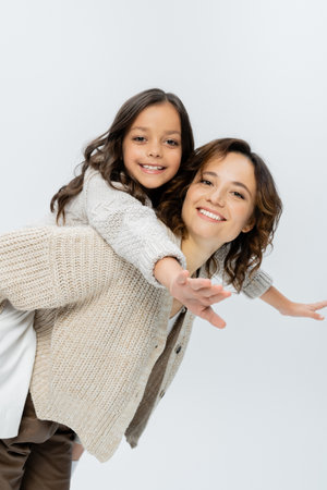 smiling woman playing with cheerful daughter while piggybacking her isolated on greyの写真素材