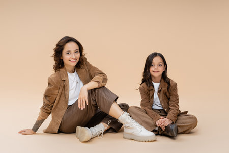 mother and daughter in fashionable autumn outfit sitting on beige backgroundの写真素材