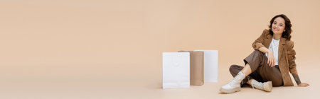 smiling woman in trendy boots and brown autumn clothes sitting near shopping bags on beige background, bannerの写真素材