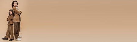 mother and daughter in suede jackets and brown pants standing back to back on beige background, bannerの写真素材