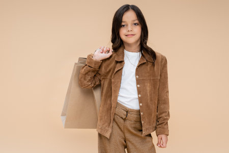 brunette girl with shopping bags posing in suede jacket and looking at camera isolated on beigeの写真素材