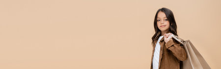 girl in fashionable autumn jacket looking at camera and holding shopping bags isolated on beige, bannerの写真素材