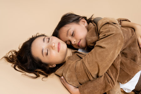 mother and child in stylish suede jackets lying with closed eyes and hugging on beige backgroundの写真素材