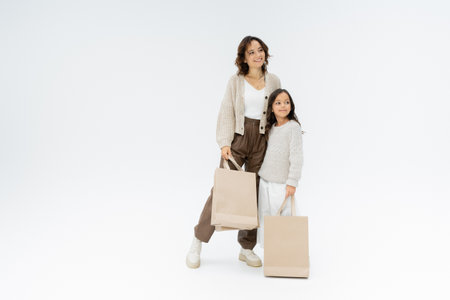 full length of trendy mother and daughter with shopping bags looking away on grey backgroundの写真素材