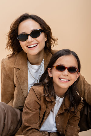 happy and stylish mother and daughter in suede jackets and sunglasses isolated on beigeの写真素材