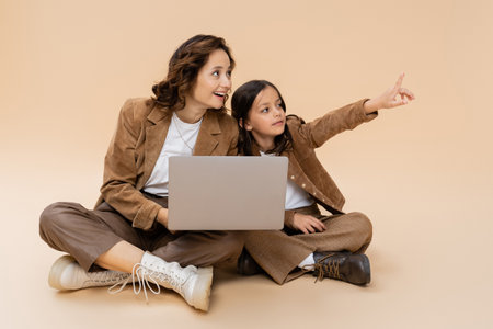 girl in trendy autumn clothes pointing with finger near laptop and smiling mom on beige backgroundの写真素材