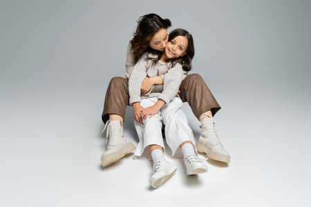 full length of stylish woman embracing daughter while sitting on grey backgroundの写真素材