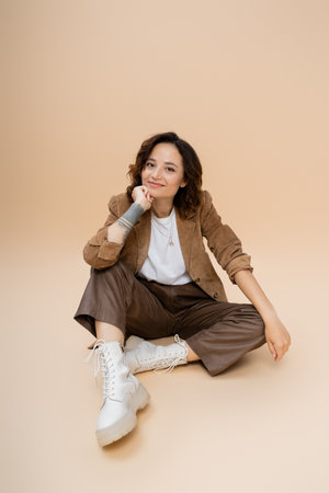 brunette woman with wavy hair wearing trendy autumn clothes while sitting with hand near face on...