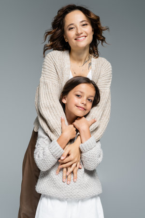 excited brunette woman in trendy knitwear hugging smiling daughter isolated on greyの写真素材