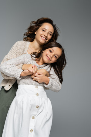 low angle view of happy young woman in knitwear hugging smiling daughter in skirt isolated on greyの写真素材