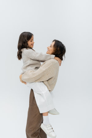 side view of brunette woman in stylish autumn clothes holding happy daughter isolated on greyの写真素材