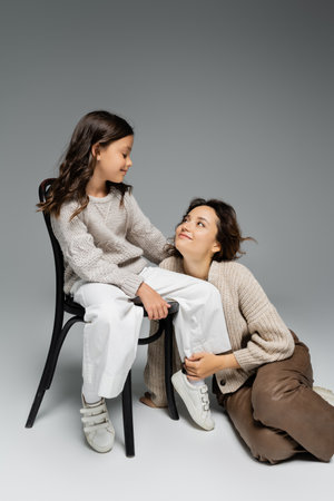 happy woman looking at daughter sitting on chair in trendy autumn outfit on grey backgroundの写真素材