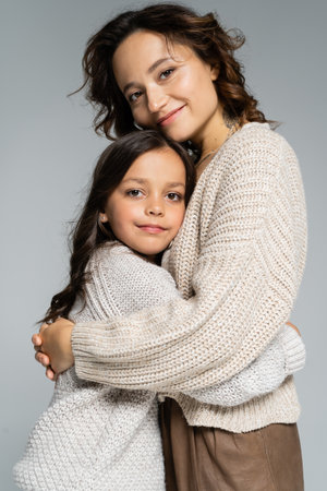 cheerful mom and daughter in cozy knitwear hugging and smiling at camera isolated on greyの写真素材