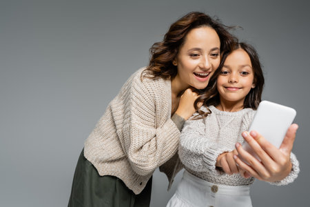smiling girl showing blurred mobile phone to amazed mom isolated on greyの写真素材