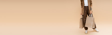 partial view of woman in brown pants and suede jacket standing with shopping bags on beige background, bannerの写真素材