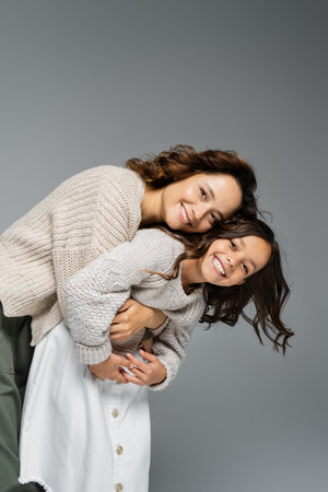 cheerful woman and child in warm outfit embracing and smiling at camera isolated on greyの写真素材