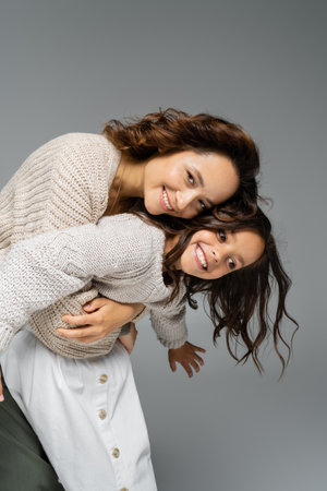 cheerful woman embracing girl in trendy autumn outfit and smiling at camera isolated on greyの写真素材