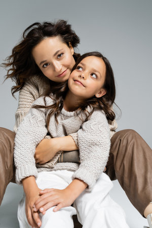 stylish and happy woman looking at camera while hugging daughter on grey backgroundの写真素材