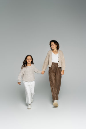 full length of mom and daughter in warm knitwear and pants walking while holding hands on grey...