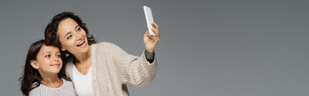 stylish and cheerful woman taking selfie with smiling daughter isolated on grey, banner