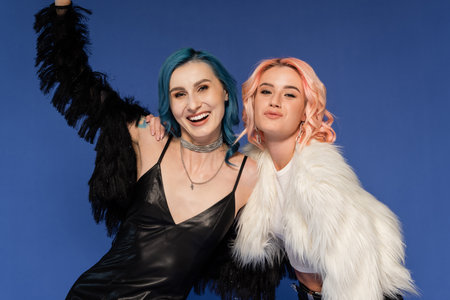 queer person hugging friend in black faux fur jacket posing with raised hand isolated on blueの写真素材