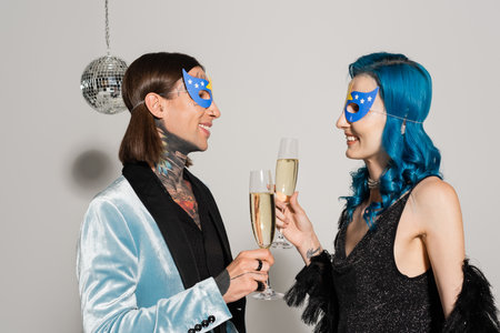side view of elegant nonbinary friends in party masks clinking champagne glasses on grey backgroundの写真素材
