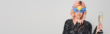 smiling woman in party mask holding champagne and showing hush sign isolated on grey, bannerの写真素材