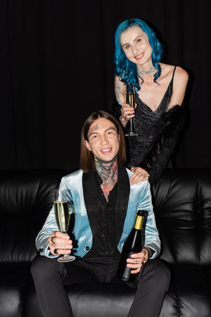 happy and elegant nonbinary friends with champagne smiling at camera on black backgroundの写真素材