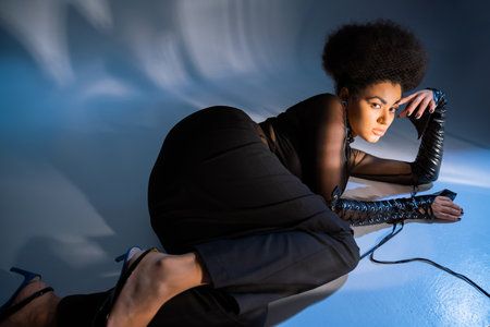 curly african american woman in transparent shirt and lace-up leather sleeves lying on blueの写真素材