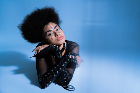 young african american model in black veil looking at camera while posing on blue backgroundの写真素材