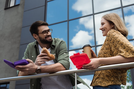 Smiling businessman holding sandwich and lunch box near colleague with coffee to go outdoorsの写真素材