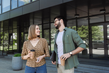 Smiling businesswoman holding coffee and cellphone near colleague with laptop outdoors