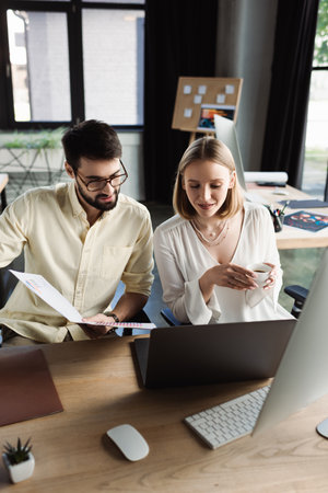 Manager holding papers near new worker with coffee cup and laptop in office