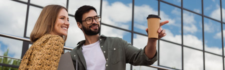 Positive businessman holding coffee to go and pointing with finger near colleague outdoors, bannerの写真素材
