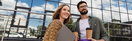 Smiling businesswoman holding paper folder near colleague with lunch box and coffee outdoors, banner