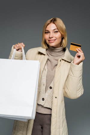 Smiling young woman in winter outfit holding shopping bags and credit card isolated on grey