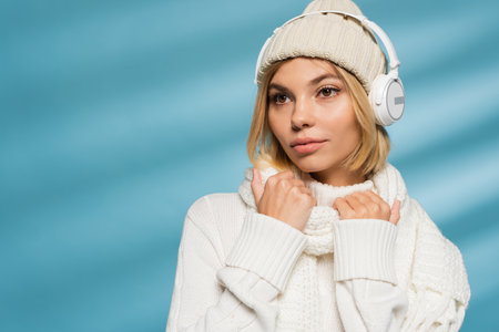 blonde young woman in knitted hat and wireless headphones looking away on blueの写真素材