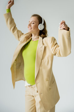 Cheerful pregnant woman in headphones and jacket standing isolated on greyの写真素材