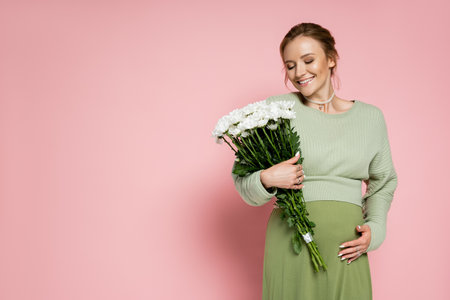 Positive pregnant woman in green outfit looking at bouquet on pink background