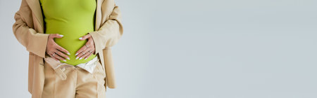 Cropped view of pregnant woman in beige suit touching belly isolated on grey, banner