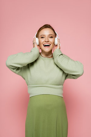 Excited pregnant woman listening music in headphones and looking at camera on pink backgroundの写真素材