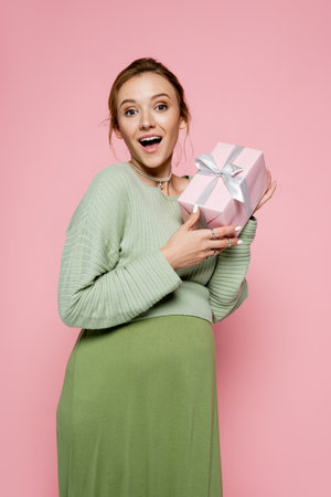 Excited pregnant woman in green outfit holding gift isolated on pinkの写真素材