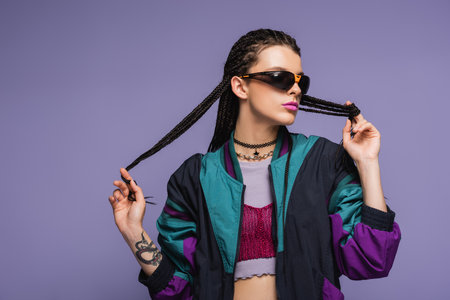 Trendy woman in retro sports jacket touching braids isolated on purpleの写真素材