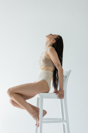 Side view of fit woman with braids sitting on chair isolated on greyの写真素材