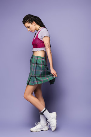 side view of tattooed woman in plaid skirt and white sneakers standing on purple backgroundの写真素材