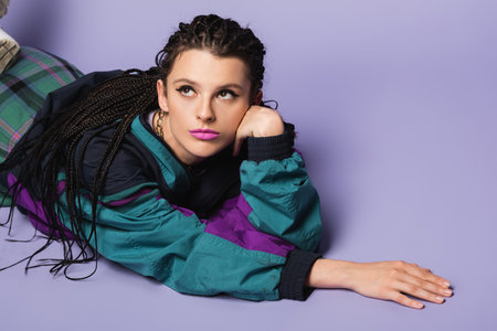 dreamy woman with braids lying in vintage style jacket and looking away on purple background