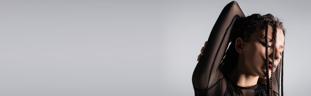 brunette woman with makeup and braids posing with closed eyes and hands behind head isolated on grey, bannerの写真素材